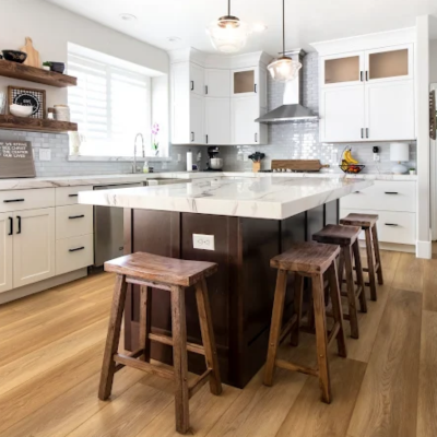Kitchen with a large center island, stainless range hood with grey backsplash tile, white cabinetry, pendant lights, built-in shelves, and several wooden bar stools in a clean, open space.